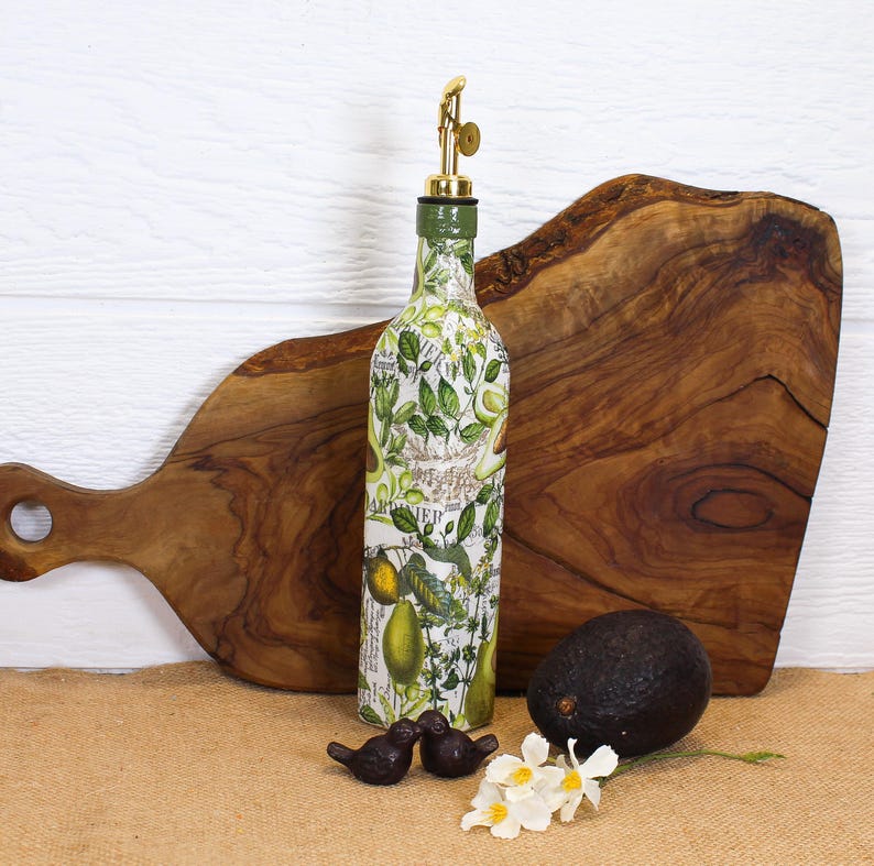 May include: A glass oil dispenser bottle decorated with a botanical print of green avocados, leaves, and text, topped with a gold-colored metal pour spout. The bottle is positioned in front of a wooden cutting board with a dark brown avocado nearby.