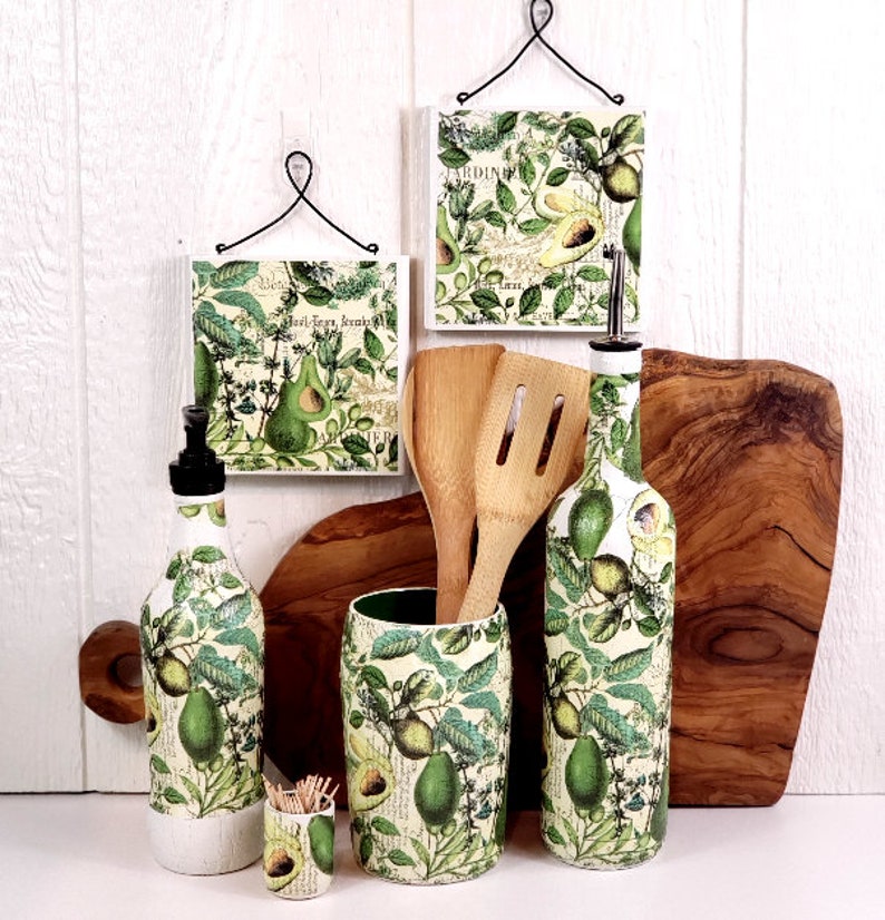 May include: A kitchen set featuring two square wall plaques with wire hangers, a soap dispenser, a utensil holder with wooden spoons, a small toothpick holder, and a tall oil bottle, all decorated with a green avocado and botanical print.