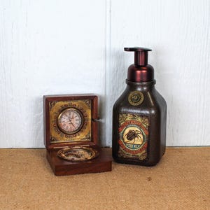 K&ouml;nnte beinhalten: Eine dunkelbraune Holzschatulle mit einer runden Uhr mit r&ouml;mischen Ziffern und einem Kompass, neben einem dunkelbraunen Seifenspender mit einem Etikett, das Doctor Kellogg Imported Spider Milk Curious Cures und eine Spinne zeigt.