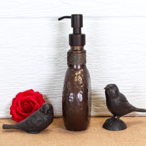 K&ouml;nnte beinhalten: Ein brauner Seifenspender aus Glas mit einem dunklen Metallpumpmechanismus und einem Seilakzent am Hals, flankiert von zwei kleinen Vogel-Figuren aus dunklem Metall und einer einzelnen leuchtend roten Rosenbl&uuml;te links vom Spender.