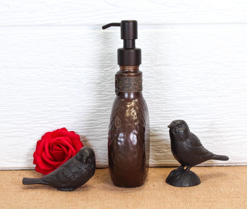 K&ouml;nnte beinhalten: Ein brauner Seifenspender aus Glas mit einem dunklen Metallpumpmechanismus und einem Seilakzent am Hals, flankiert von zwei kleinen Vogel-Figuren aus dunklem Metall und einer einzelnen leuchtend roten Rosenbl&uuml;te links vom Spender.