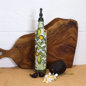 May include: A tall glass bottle with a black pourer spout, decorated with a botanical print of yellow lemons and green leaves on a white background. The bottle is positioned upright, featuring a cylindrical shape and a tapered neck design.
