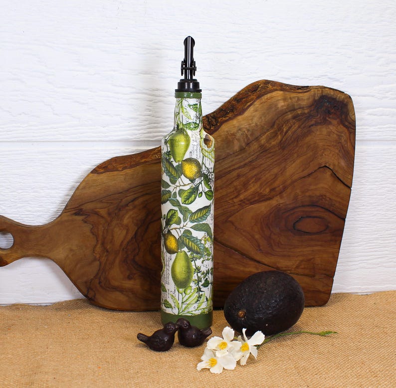 May include: A tall glass bottle with a black pourer spout, decorated with a botanical print of yellow lemons and green leaves on a white background. The bottle is positioned upright, featuring a cylindrical shape and a tapered neck design.