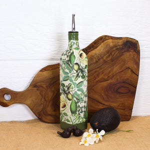 May include: A glass oil dispenser bottle featuring a botanical print of green avocados and leaves on a cream background, topped with a silver metal pour spout. The base and neck of the bottle are painted in a solid dark olive green color.