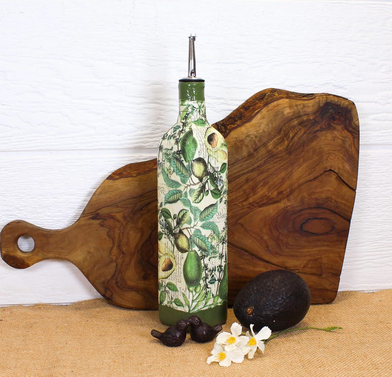 May include: A glass oil dispenser bottle featuring a botanical print of green avocados and leaves on a cream background, topped with a silver metal pour spout. The base and neck of the bottle are painted in a solid dark olive green color.