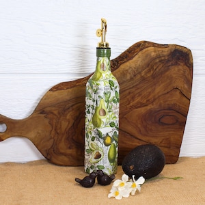 May include: A glass oil dispenser bottle featuring a decorative print of green avocados and botanical leaves. The bottle is topped with a gold-colored metal pour spout and a green cap. The design includes subtle text elements integrated into the pattern.