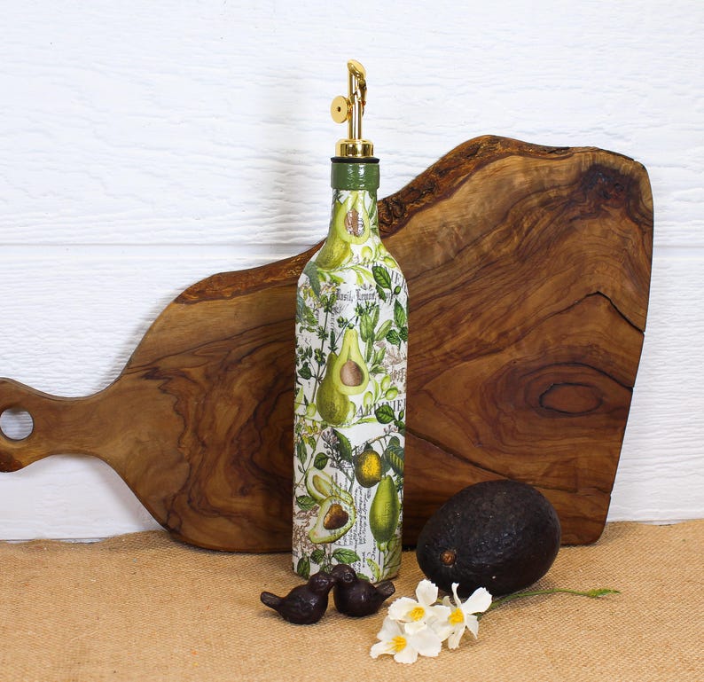 May include: A glass oil dispenser bottle featuring a decorative print of green avocados and botanical leaves. The bottle is topped with a gold-colored metal pour spout and a green cap. The design includes subtle text elements integrated into the pattern.