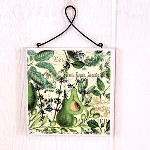 May include: A square decorative tile featuring a botanical illustration of avocados and herbs on a cream background. The design includes the text Basil, Lemon, Avocado and Botanique. A black metal wire hanger is attached to the top of the tile.