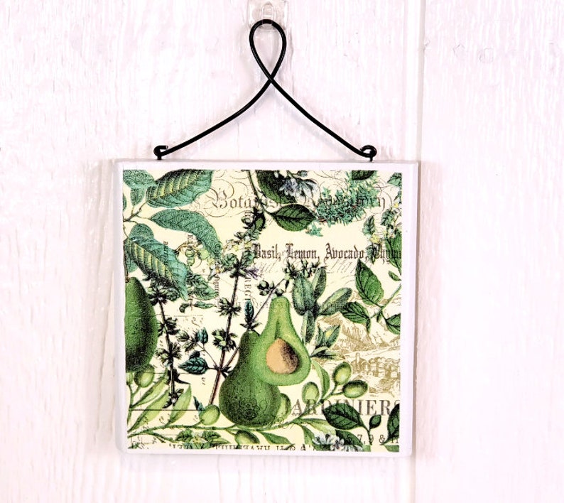 May include: A square decorative tile featuring a botanical illustration of avocados and herbs on a cream background. The design includes the text Basil, Lemon, Avocado and Botanique. A black metal wire hanger is attached to the top of the tile.