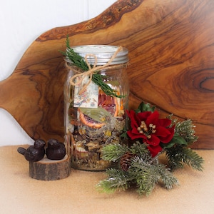 Op de afbeelding: Een glazen pot gevuld met gedroogde botanische producten, vastgebonden met touw en groen. Een rode poinsettia en decoratieve vogels op een houten voet staan op de voorgrond. De pot staat op een jute oppervlak met een houten snijplank.