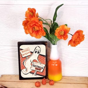 Op de afbeelding: Een houten bord met een spook dat een bord vasthoudt met de tekst "Alstublieft geen grappen" en "Hier is je Halloween-traktatie". Het bord is versierd met oranje en zwarte stippen. Het bord staat op een houten tafel met een vaas met oranje bloemen en kleine oranje pompoenen.