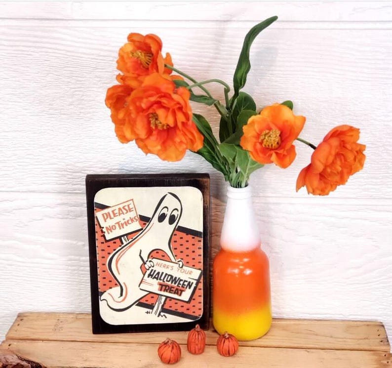 Op de afbeelding: Een houten bord met een spook dat een bord vasthoudt met de tekst "Alstublieft geen grappen" en "Hier is je Halloween-traktatie". Het bord is versierd met oranje en zwarte stippen. Het bord staat op een houten tafel met een vaas met oranje bloemen en kleine oranje pompoenen.