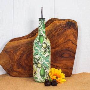 May include: A glass oil dispenser bottle featuring a botanical print of green avocados and leaves on a cream background. The bottle is topped with a silver-colored metal pour spout. The design includes detailed illustrations of whole and sliced avocados.