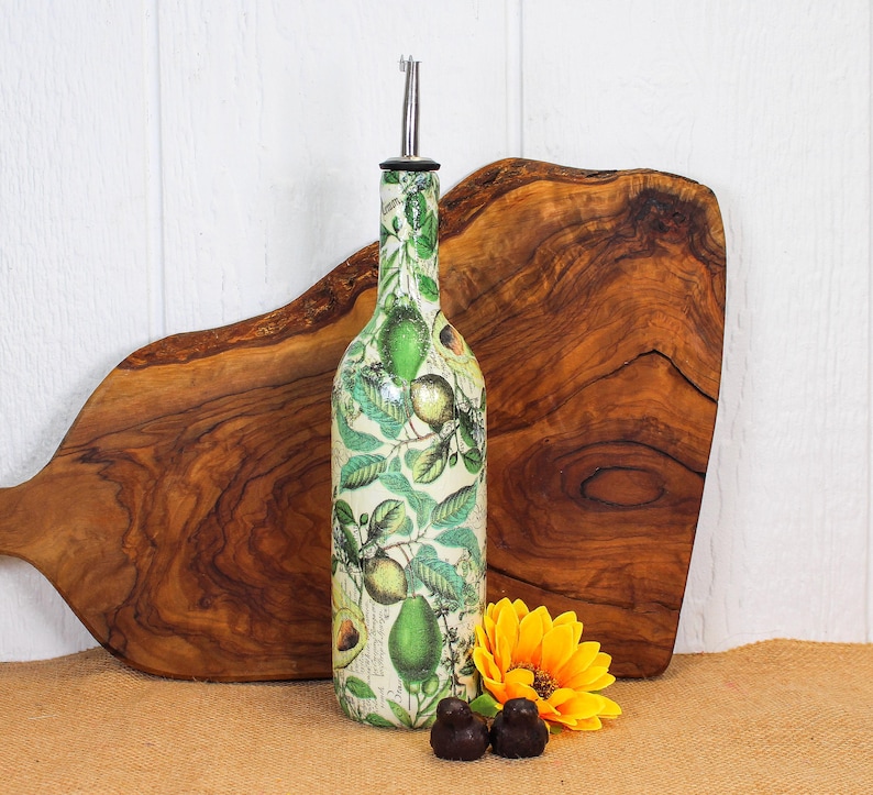 May include: A glass oil dispenser bottle featuring a botanical print of green avocados and leaves on a cream background. The bottle is topped with a silver-colored metal pour spout. The design includes detailed illustrations of whole and sliced avocados.