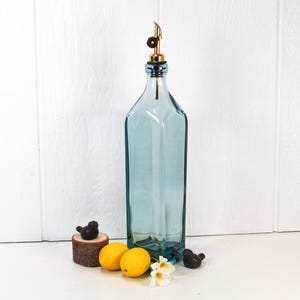 May include: A tall, clear blue glass bottle with a gold metal spout and stopper. The bottle is sitting on a white surface with two lemons and a small bird figurine.