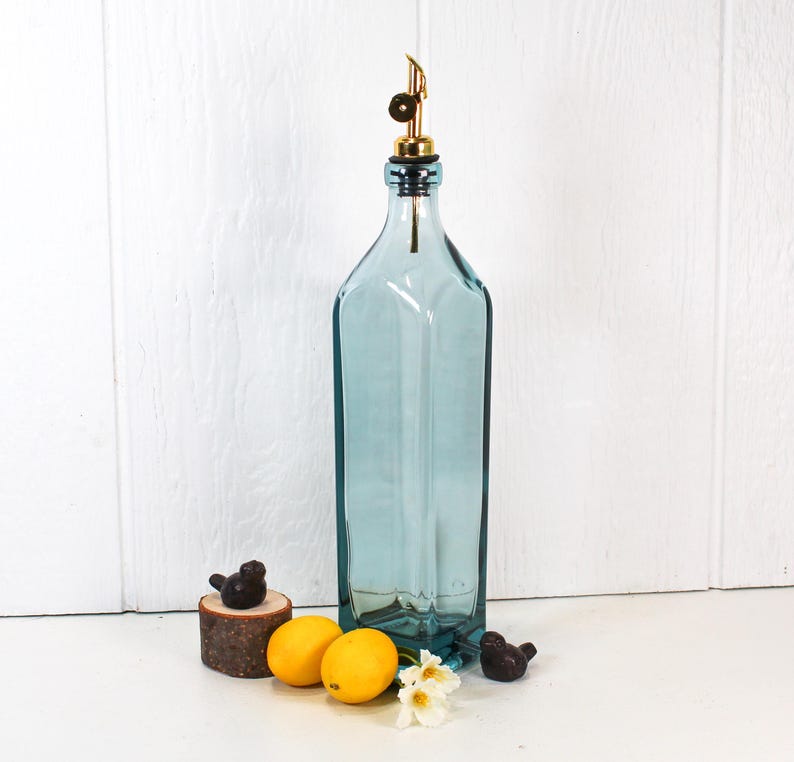 May include: A tall, clear blue glass bottle with a gold metal spout and stopper. The bottle is sitting on a white surface with two lemons and a small bird figurine.