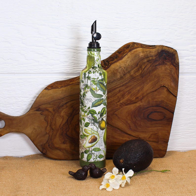 May include: A tall glass bottle with a black pour spout, decorated with a botanical print featuring green avocado illustrations and leaves. The bottle is positioned upright, accompanied by a whole dark avocado and two small bird figurines.