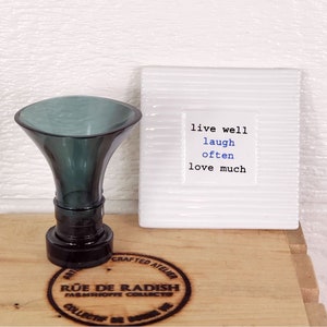 Pu&ograve; includere: Un tappo per bottiglia in vetro verde scuro con una base rotonda e una parte superiore larga a forma di imbuto. Il tappo &egrave; su una superficie di legno con un logo circolare che dice "Rue de Radish Farmshoppe Collectibles". Sullo sfondo c'&egrave; una piastrella di ceramica bianca con il testo "live well laugh often love much".