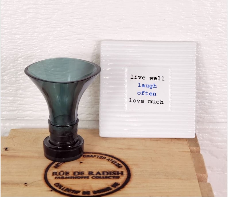 Pu&ograve; includere: Un tappo per bottiglia in vetro verde scuro con una base rotonda e una parte superiore larga a forma di imbuto. Il tappo &egrave; su una superficie di legno con un logo circolare che dice "Rue de Radish Farmshoppe Collectibles". Sullo sfondo c'&egrave; una piastrella di ceramica bianca con il testo "live well laugh often love much".
