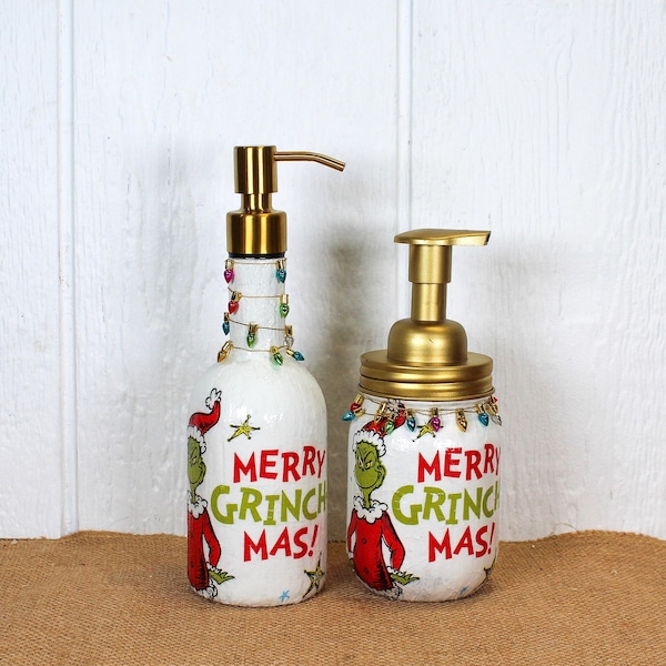 Pode incluir: Dois dispensadores de sabonete brancos com bombas douradas, cada um com um personagem Grinch verde em um traje vermelho e o texto MERRY GRINCH MAS em vermelho e verde. Ambas as garrafas são decoradas com um fio de mini luzes de Natal coloridas.