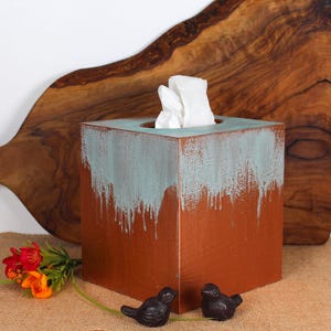 May include: A square tissue box with a distressed copper finish and teal paint drips. The box holds white tissues and is decorated with two small black bird figurines. A wooden cutting board is in the background.