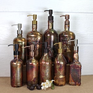 May include: A collection of nine glass soap dispensers with a distressed, metallic finish in shades of bronze, copper, and gold. Each bottle features a pump mechanism in varying metallic tones, including matte black, brushed silver, and polished gold.
