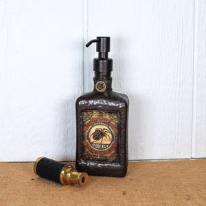 K&ouml;nnte beinhalten: Ein dunkelbrauner, texturierter Seifenspender mit schwarzem Pumpmechanismus und einem Etikett im Vintage-Stil mit der Aufschrift Doctor Kellogg's Imported Spider Milk Curious Cures. Daneben liegt ein kleines zylindrisches Objekt aus Messing und Schwarz.