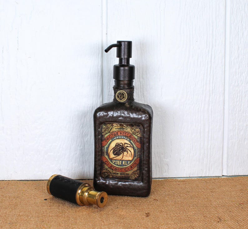 K&ouml;nnte beinhalten: Ein dunkelbrauner, texturierter Seifenspender mit schwarzem Pumpmechanismus und einem Etikett im Vintage-Stil mit der Aufschrift Doctor Kellogg's Imported Spider Milk Curious Cures. Daneben liegt ein kleines zylindrisches Objekt aus Messing und Schwarz.