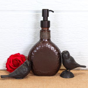 K&ouml;nnte beinhalten: Ein dunkelbrauner Seifenspender mit geh&auml;mmerter Textur und Pumpmechanismus, der am Hals mit einer gewickelten Kordel verziert ist. Der Spender wird von zwei kleinen schwarzen Vogelfiguren und einer einzelnen roten Rosenbl&uuml;te flankiert.