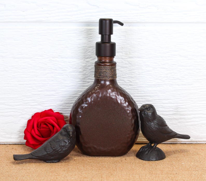 K&ouml;nnte beinhalten: Ein dunkelbrauner Seifenspender mit geh&auml;mmerter Textur und Pumpmechanismus, der am Hals mit einer gewickelten Kordel verziert ist. Der Spender wird von zwei kleinen schwarzen Vogelfiguren und einer einzelnen roten Rosenbl&uuml;te flankiert.