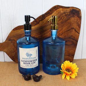 May include: Two blue glass bottles with black and gold pump dispensers. One bottle has a label that reads "Drumshanbo Gunpowder Irish Gin." Two small, dark bird figurines and a yellow sunflower are in front of the bottles.
