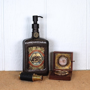 K&ouml;nnte beinhalten: Ein dunkelbrauner, texturierter Seifenspender mit schwarzer Pumpe und einem Etikett im Vintage-Stil mit der Aufschrift Doctor Kellogg's Snake Oil. Daneben liegen ein Fernrohr aus Messing und schwarzem Leder sowie eine kleine Holzschatulle mit Uhr.