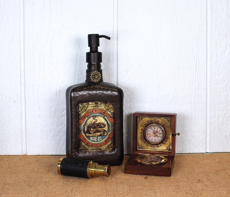K&ouml;nnte beinhalten: Ein dunkelbrauner, texturierter Seifenspender mit schwarzer Pumpe und einem Etikett im Vintage-Stil mit der Aufschrift Doctor Kellogg's Snake Oil. Daneben liegen ein Fernrohr aus Messing und schwarzem Leder sowie eine kleine Holzschatulle mit Uhr.