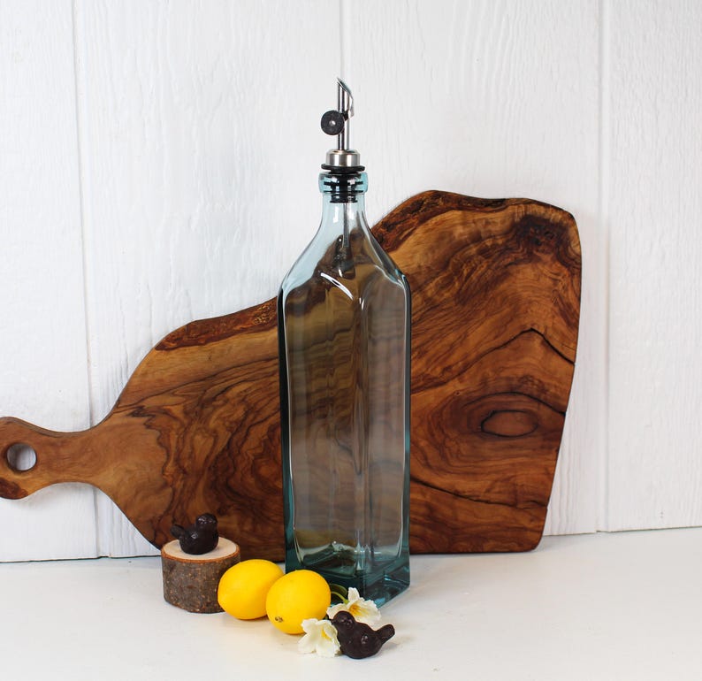 May include: A clear glass bottle with a silver spout and a blue tint. The bottle is sitting on a wooden cutting board. There are two small bird figurines and lemons in the foreground.