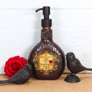 K&ouml;nnte beinhalten: Ein dunkelbrauner, geh&auml;mmerter Seifenspender mit Pumpkopf und einem Etikett mit der Aufschrift PREMIUM QUALITY BLOOD OF VAMPIRES. Die Flasche wird von einer roten Rose und zwei kleinen schwarzen Vogelfiguren als Dekoration flankiert.