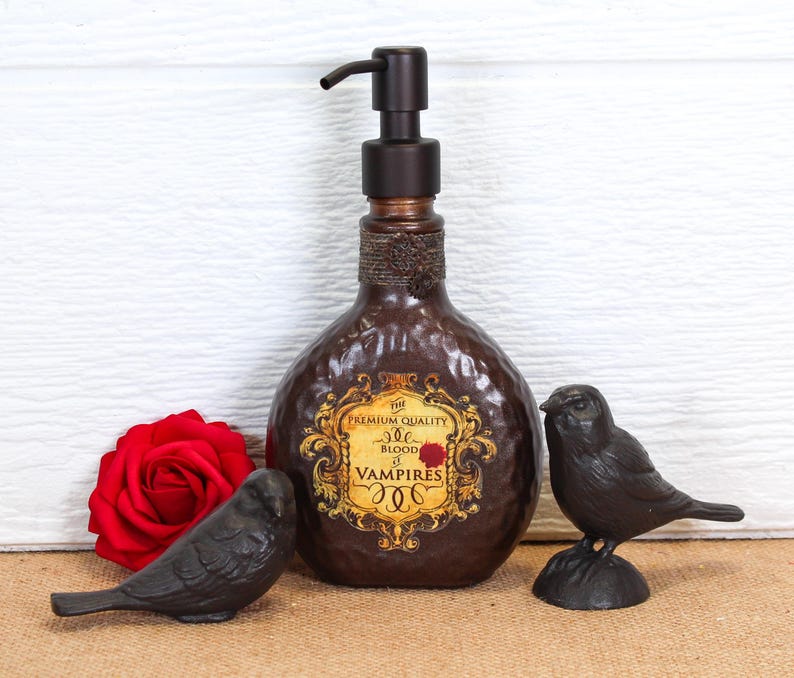 K&ouml;nnte beinhalten: Ein dunkelbrauner, geh&auml;mmerter Seifenspender mit Pumpkopf und einem Etikett mit der Aufschrift PREMIUM QUALITY BLOOD OF VAMPIRES. Die Flasche wird von einer roten Rose und zwei kleinen schwarzen Vogelfiguren als Dekoration flankiert.