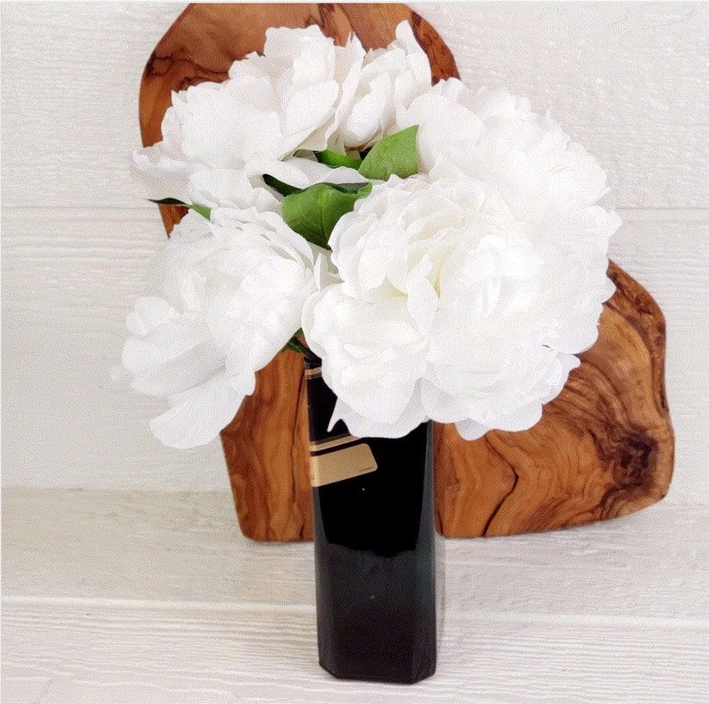 May include: A bouquet of white peonies arranged in a black and gold vase. The vase is sitting on a wooden heart-shaped cutting board.