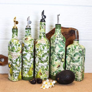 May include: Five glass bottles with pour spouts, decorated with a green avocado and botanical print. The bottles vary in height and shape, featuring metallic or black stoppers. A single dark avocado and small white flowers sit in the foreground.