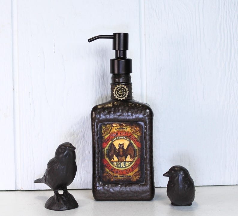 K&ouml;nnte beinhalten: Ein dunkelbrauner, texturierter rechteckiger Seifenspender mit schwarzer Pumpe und einem Etikett mit Fledermaus-Illustration und dem Text Doctor Kellogg's Imported Bat's Blood Curious Cure. Zwei kleine, dunkle Vogelfiguren stehen daneben.
