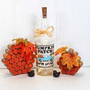 May include: A clear glass bottle with a cork top and a label that reads "Pumpkin Patch Farm Grown" with a blue truck and pumpkins. The bottle is decorated with a brown burlap bow and surrounded by two stacks of cork rounds with fall leaves.