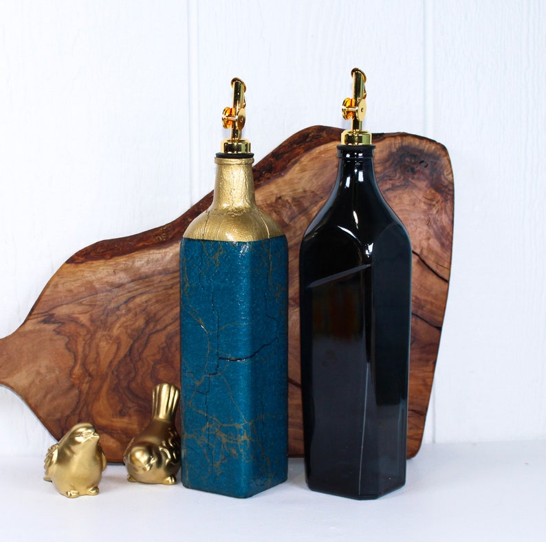May include: Two glass bottles with gold tops and spouts, one painted teal with gold accents and the other black, sitting on a wooden chopping board.