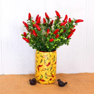 May include: A yellow ceramic vase with a red pepper pattern, filled with a bouquet of red pepper stems and green leaves. The vase is sitting on a brown burlap surface.