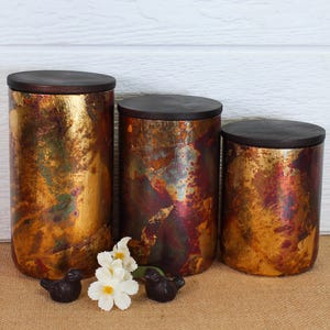 May include: Three cylindrical canisters of varying heights with dark brown wooden lids. The canisters feature a metallic finish with mottled patches of gold, copper, and deep red. Two small dark bird figurines and a white flower sit in front.