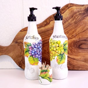 May include: Two white, decorative condiment dispensers with black pumps, labeled "Ketchup" and "Mustard." Each bottle features a grape design. A small matching toothpick holder is also present, with a grape leaf design.