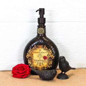 K&ouml;nnte beinhalten: Ein dunkelbrauner Seifenspender mit schwarzer Pumpe und einem dekorativen Etikett mit der Aufschrift The Premium Quality Blood of Vampires. Die Flasche wird von einer roten Rose und zwei kleinen schwarzen Vogelfiguren begleitet.