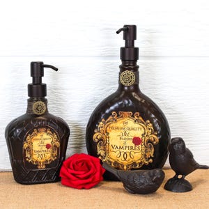 K&ouml;nnte beinhalten: Zwei dunkelbraune Seifenspender aus Glas mit schwarzen Pumpk&ouml;pfen, jeweils mit einem gold-schwarzen Etikett mit der Aufschrift The Premium Quality Blood of Vampires. Eine rote Rose und zwei kleine schwarze Vogelfiguren stehen davor.