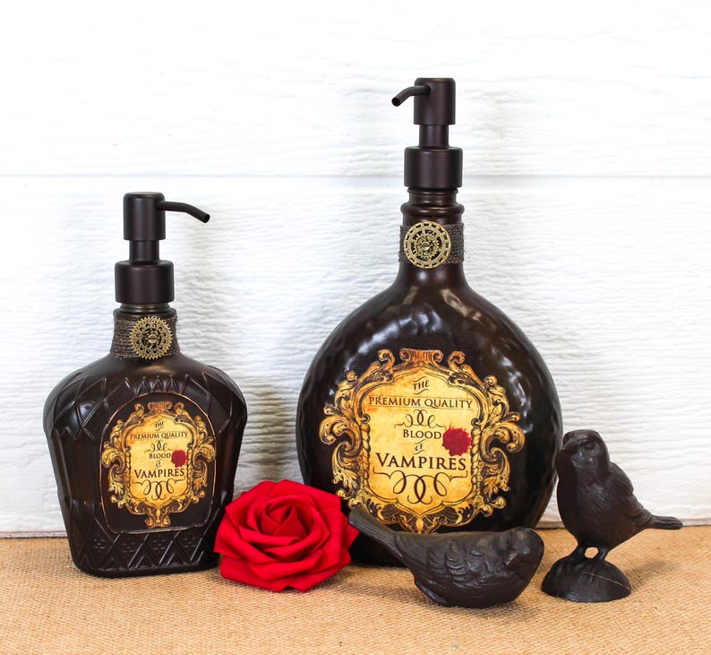 K&ouml;nnte beinhalten: Zwei dunkelbraune Seifenspender aus Glas mit schwarzen Pumpk&ouml;pfen, jeweils mit einem gold-schwarzen Etikett mit der Aufschrift The Premium Quality Blood of Vampires. Eine rote Rose und zwei kleine schwarze Vogelfiguren stehen davor.