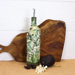 May include: A tall, cylindrical glass bottle with a metal pour spout, decorated with a botanical print featuring green avocado illustrations and script text. The bottle has a solid green base and is positioned upright with a dark green avocado.