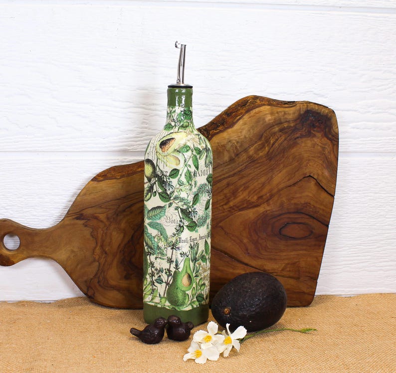 May include: A tall, cylindrical glass bottle with a metal pour spout, decorated with a botanical print featuring green avocado illustrations and script text. The bottle has a solid green base and is positioned upright with a dark green avocado.