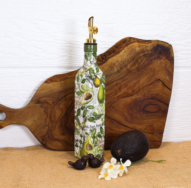 May include: A glass bottle with a gold-colored metal pourer spout, decorated with a botanical print featuring green avocados and leaves on a white background. The bottle is positioned upright, accompanied by a whole dark avocado and small white flowers.